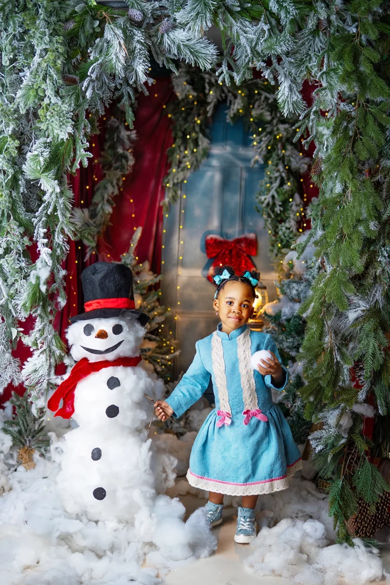 Young girl in a blue dress standing beside a snowman during a Christmas photography session in Enfield, North London
