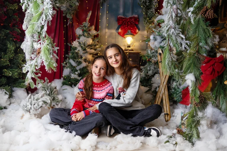 Two sisters sitting together in a snowy Christmas studio setup during a photography session in Enfield, North London