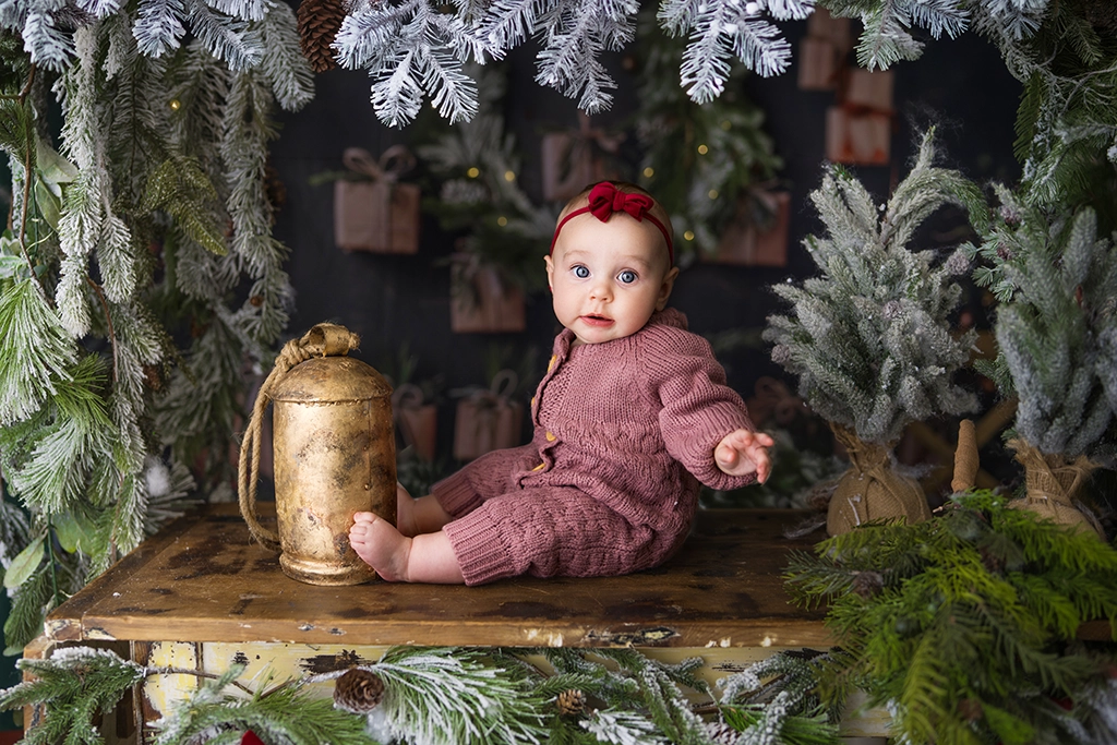 Baby wearing a mauve knitted outfit and red bow sitting beside a golden bell among frosted Christmas trees.