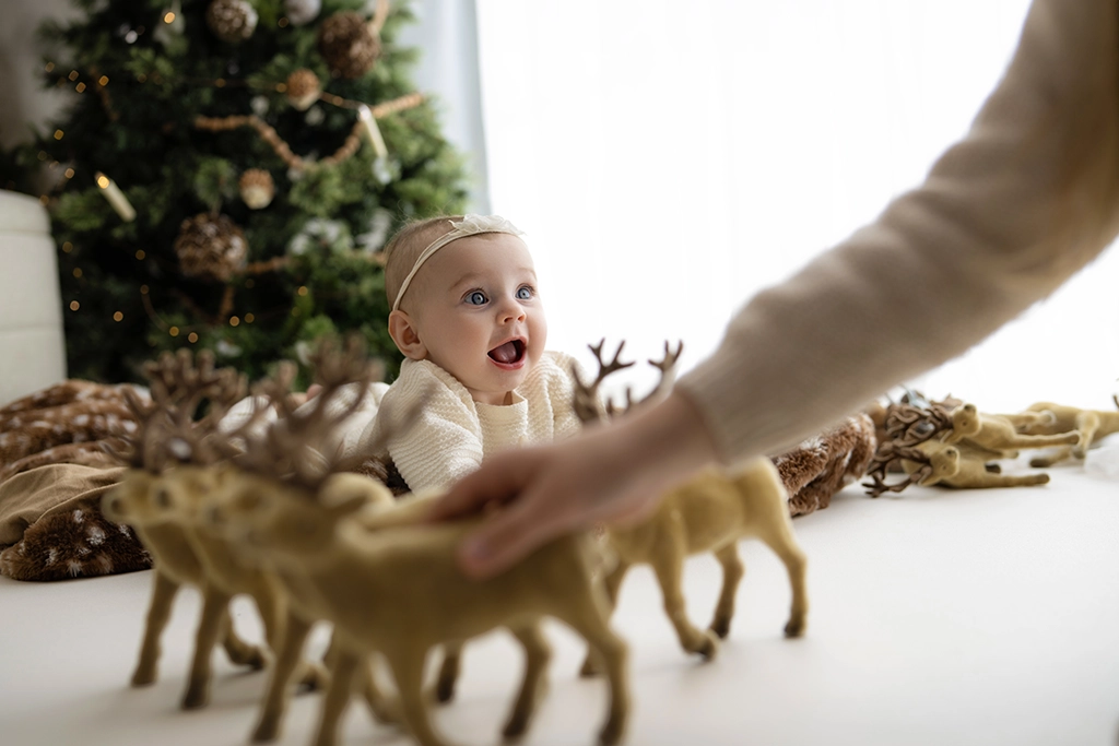 Baby girl in cream knitwear watching decorative reindeer being moved in front of her, with a Christmas tree behind her in an Enfield studio.