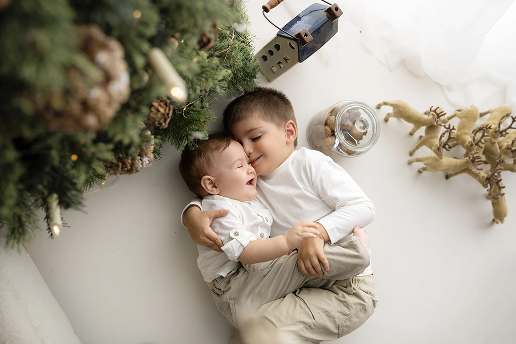 Overhead view of an older brother cuddling his baby brother beside a Christmas tree during a festive studio session in Enfield.