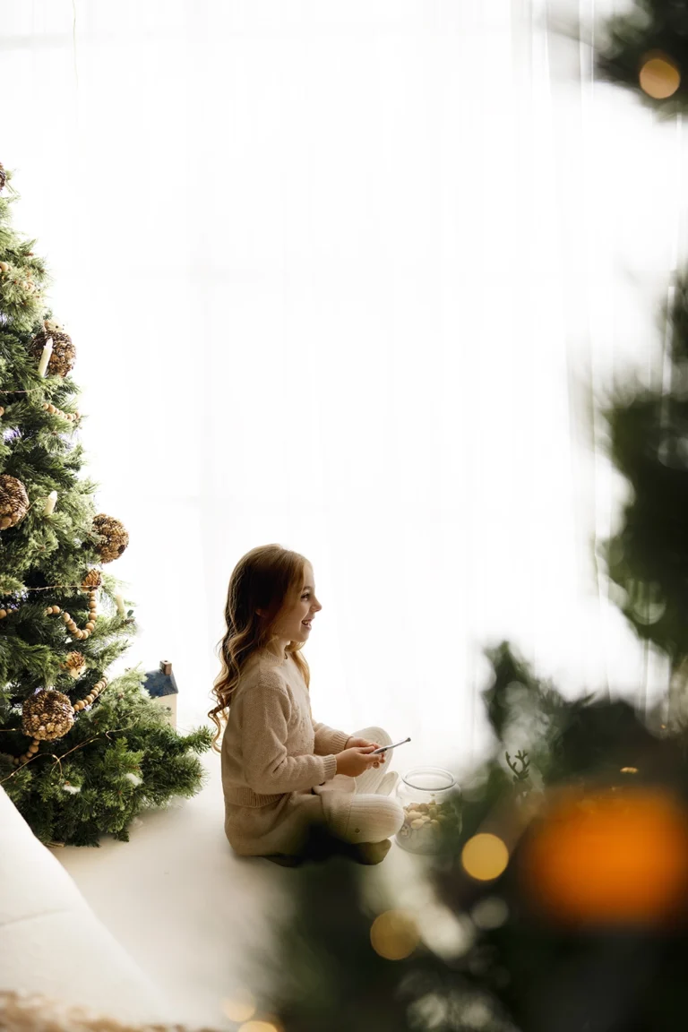 Smiling girl in cream knitwear sitting beside a Christmas tree and a bright window, framed by blurred greenery and golden lights in an Enfield studio.