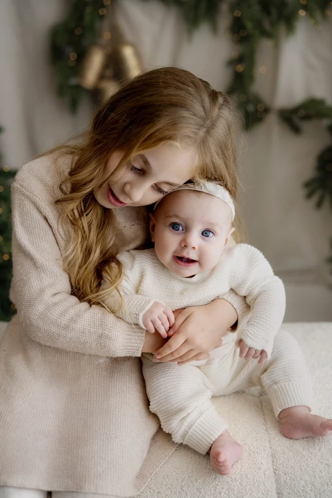 Older sister cuddling her baby sister, both wearing cream knitwear, with Christmas greenery and warm fairy lights behind them in an Enfield studio.
